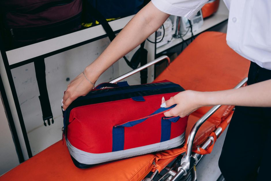 A healthcare worker reaches for a red medical bag on a stretcher indoors.